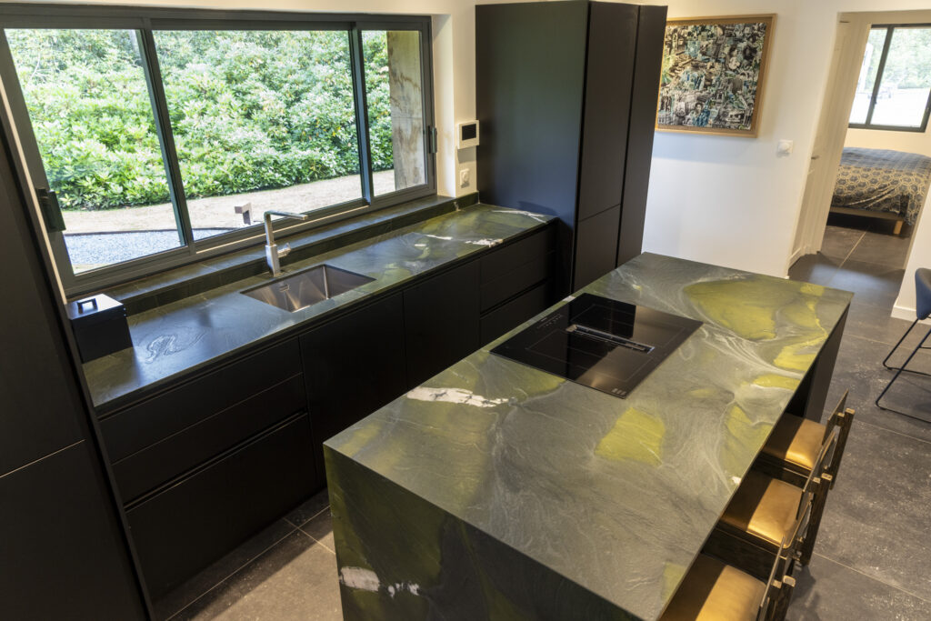 Îlot de cuisine en pierre naturelle vert et or veinée avec une plaque de cuisson intégrée, entre meubles noirs mats