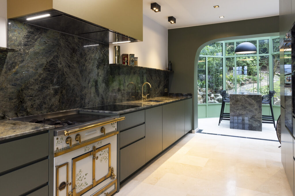 Cuisine avec îlot central et plan de travail en pierre sombre aux reflets bleutés, entourée de meubles verts et d'une porte cintrée ouvrant sur le jardin