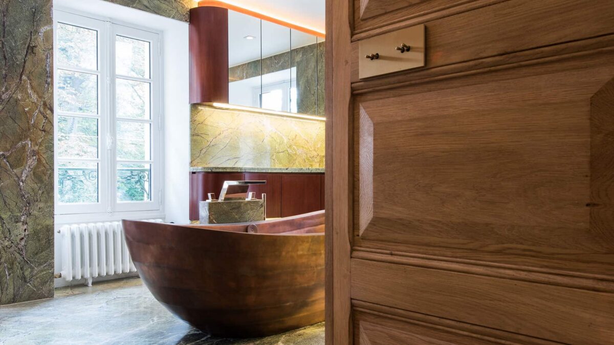Baignoire en cuivre dans une salle de bain aux murs de marbre vert, porte en bois massif et fenêtre