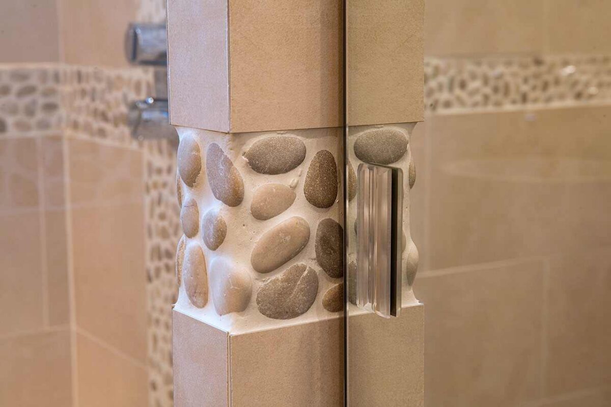 Pilier habillé de galets et parois en céramique effet pierre beige dans une salle de bain, poignée de porte de douche en verre au premier plan