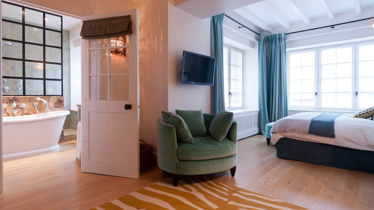 Chambre avec fauteuil vert et vue sur une salle de bain au mur de carreaux façon zellige vert d'eau et baignoire îlot
