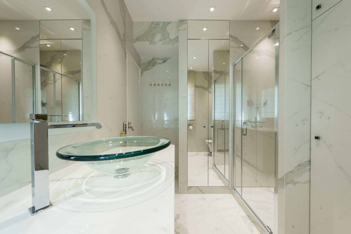 Salle de bain en céramique effet marbre blanc veiné de gris, vasque ronde en verre transparent et parois de douche en miroir