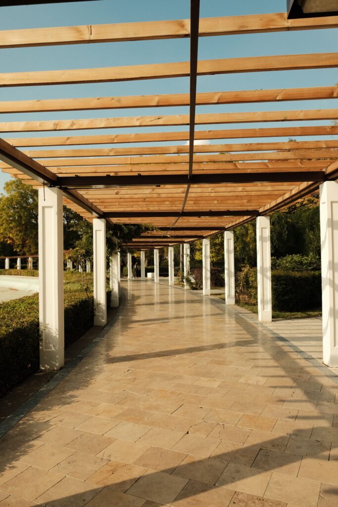 Allée en dalles de pierre beige sous une pergola en bois, bordée de colonnes claires