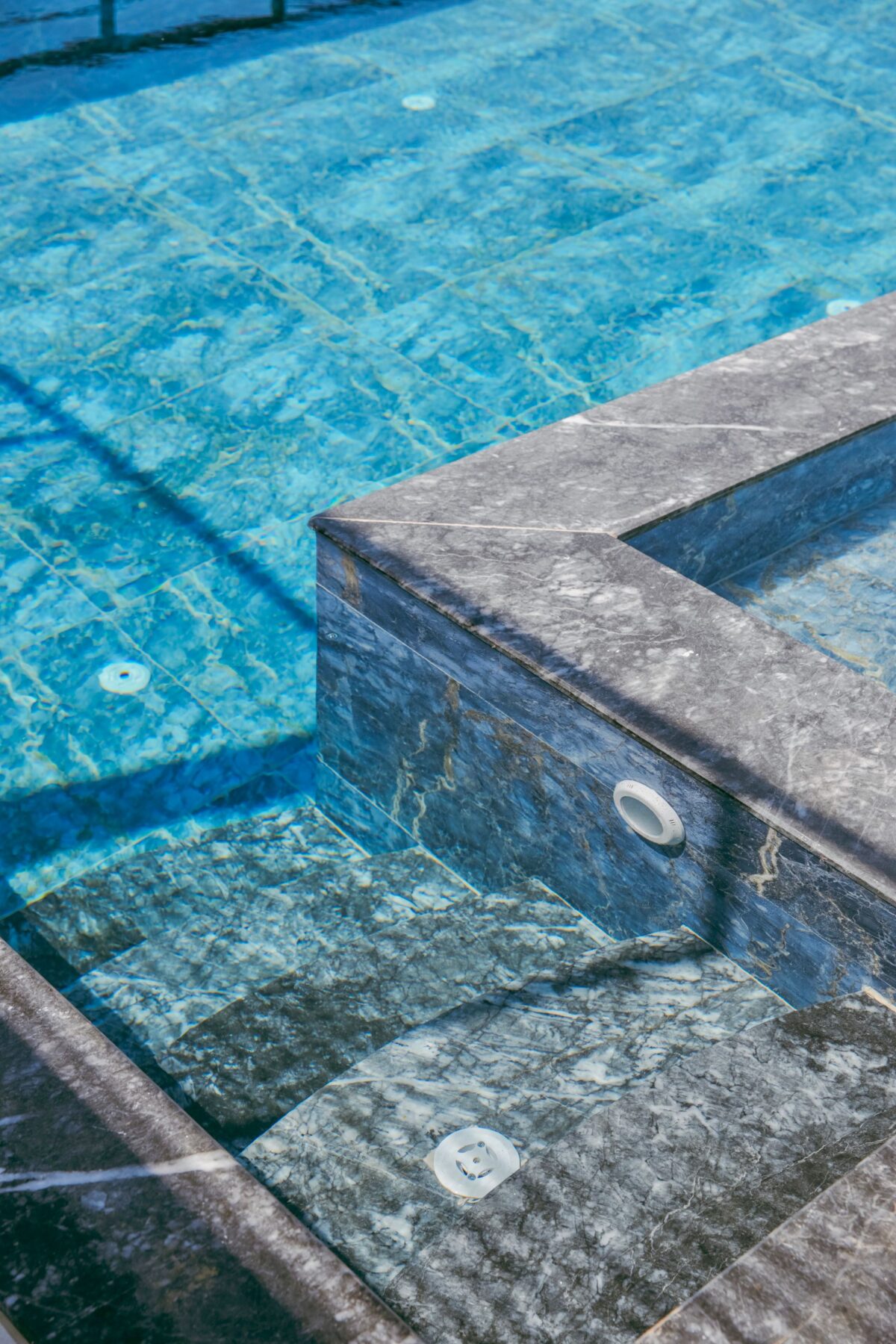 Angle de margelle et de marches en pierre grise veinée au bord d'un bassin turquoise, spots encastrés