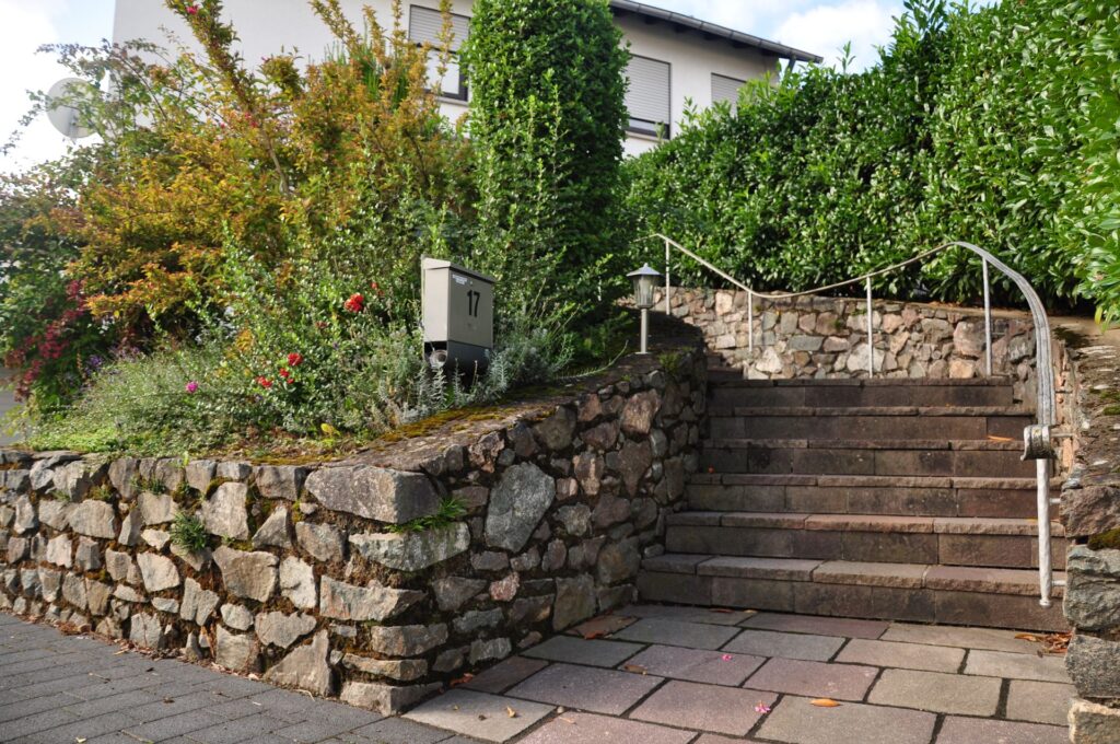 Escalier en pierre claire bordé d'un muret de pierres et de buissons, dallage au pied