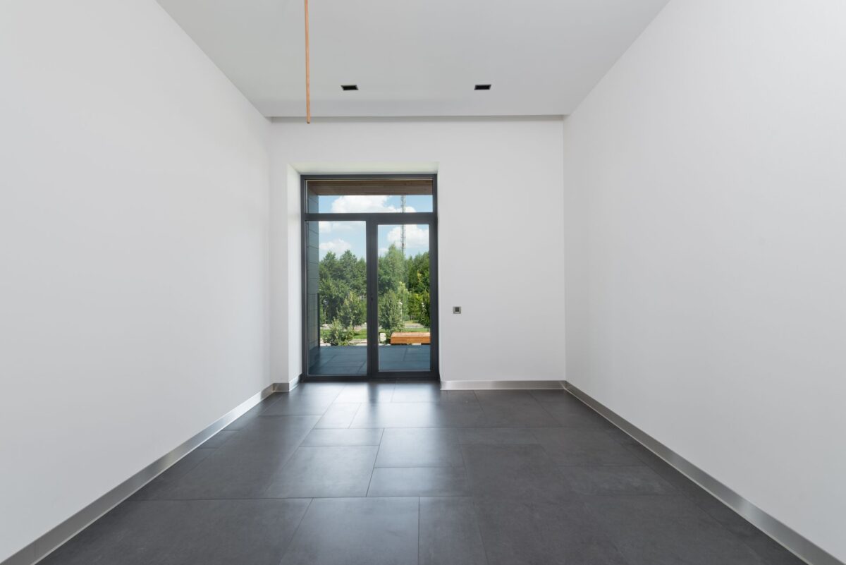 Pièce vide au sol en carrelage gris clair grand format, avec une baie vitrée ouvrant sur un jardin