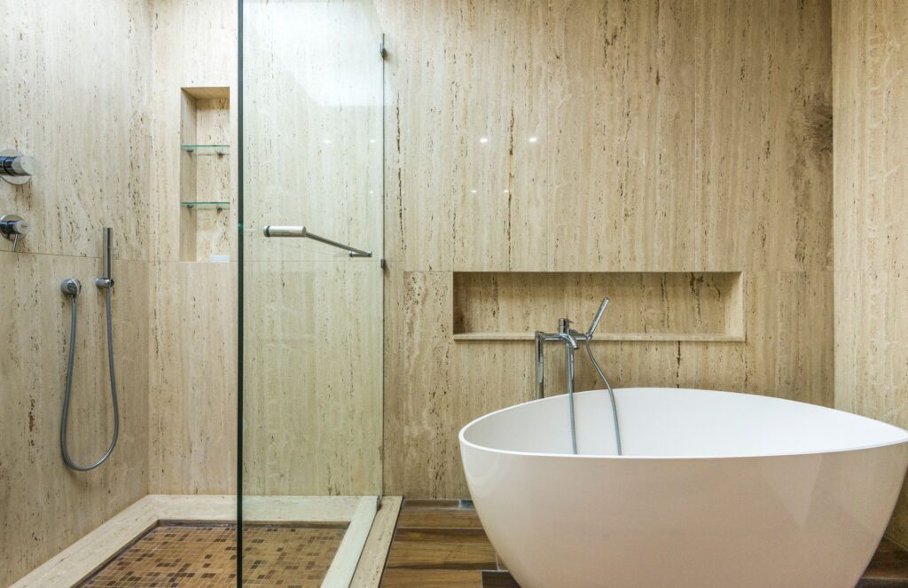 Salle de bain aux murs en travertin beige veiné, douche à l'italienne vitrée et baignoire îlot blanche